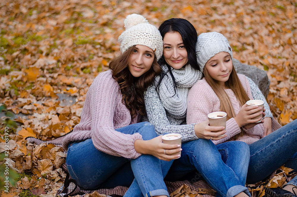 Cute family in a autumn park. Happy mother with two daughters wearing sweaters and jeans, drinking warm coffee or cocoa. Family sitting on yellow leaves. Golden autumn. Cute family in a autumn park. Happy mother with two daughters wearing sweaters and jeans, drinking warm coffee or cocoa. Family sitting on yellow leaves. Golden autumn.