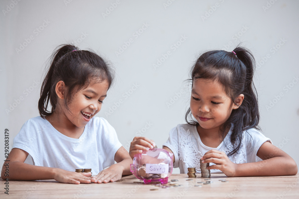Two cute asian child girls putting money into piggy bank to save money for the future together with fun and happiness Two cute asian child girls putting money into piggy bank to save money for the future together with fun and happiness