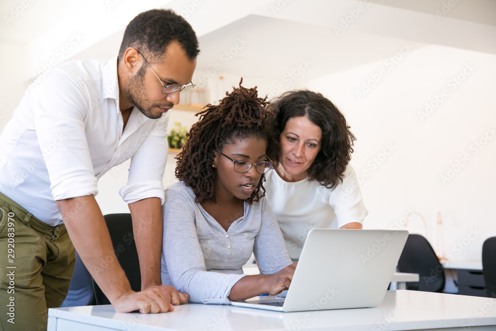 © Mangostar - Multiethnic team collaborating on project at one workplace. Business man and women in casual sitting and standing at desk, using laptop, looking at screen and talking. Collaboration concept