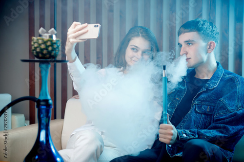 Beautiful young woman with guy hookah, blow smoke from tobacco and take selfie photo on phone, blue shisha background