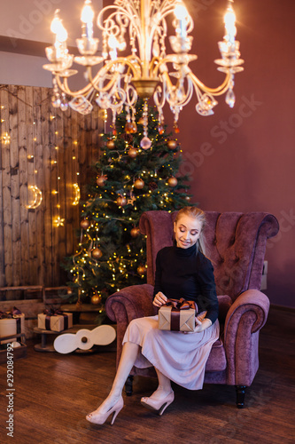 Beautiful young woman dressed in a sweater and skirt sitting on the arm chair next to the christmas tree with gift box in hands.