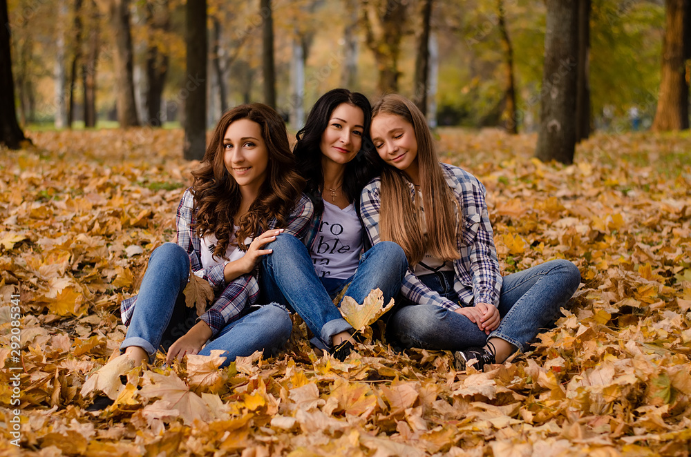 Cute family in a autumn park. Happy mother with two daughters wearing checkered shirts and jeans having fun. Family sitting on yellow leaves. Golden autumn.