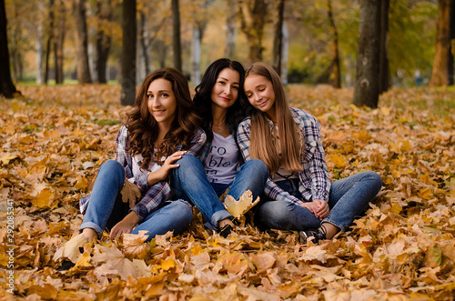 Cute family in a autumn park. Happy mother with two daughters wearing checkered shirts and jeans having fun. Family sitting on yellow leaves. Golden autumn.