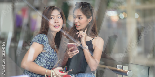 Two asian women chatting together and holding smartphone while shopping at the mall