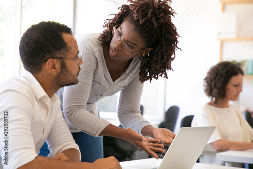 Trainer explaining software specifics to new employee. Man and woman in casual sitting and standing at desk, using laptop, pointing at screen and talking. Corporate education concept photo