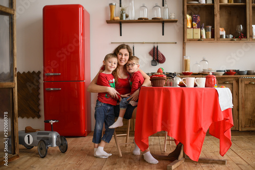 Mom, sitting at dining table, hugs two sons, in kitchen made of wood, against  background of red refrigerator