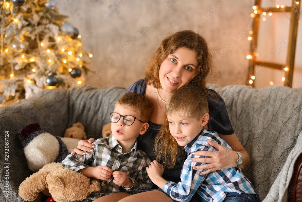 Happy and loving mom hugs her children sitting on the sofa in room, against background of Christmas tree, glowing with lights and bokeh