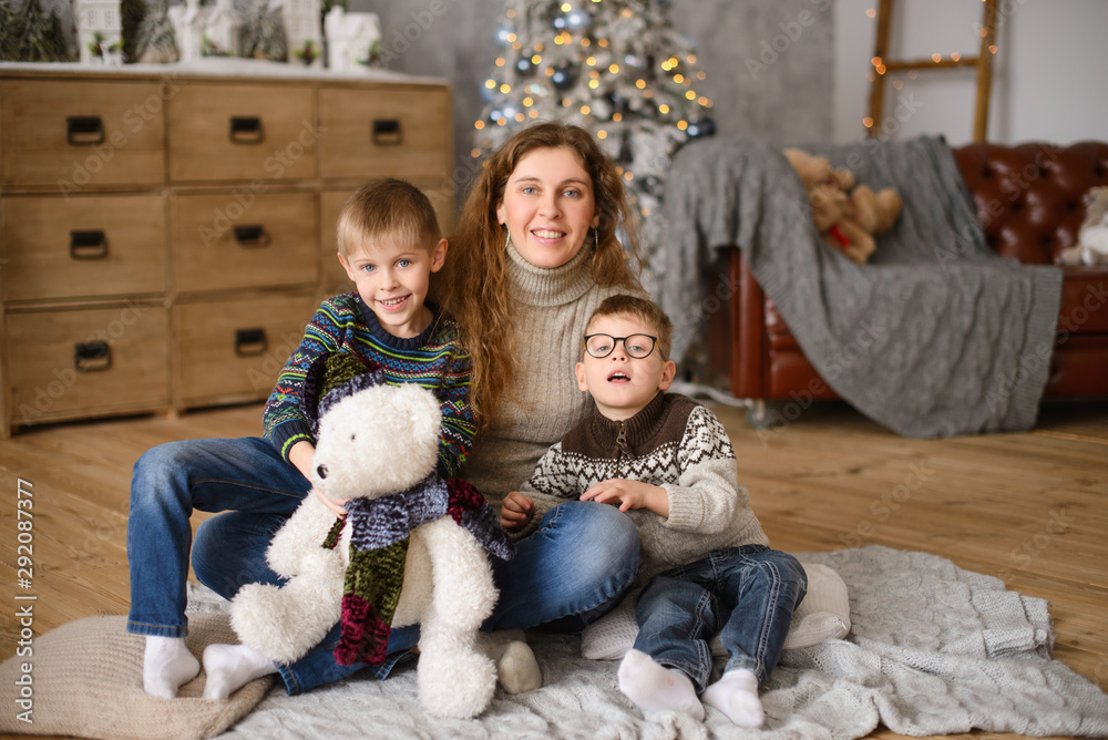 Mom with two sons in sweaters, hugging, sitting on wooden floor, in room with  Christmas tree, next to toy bear