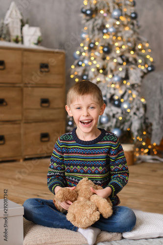 boy in sweaters, sitting on wooden floor, in room with  Christmas tree, plaing to toy bear photo