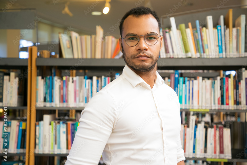 Cheerful African American man posing at public library. Front view of confident young guy standing in front of bookshelves. Knowledge concept