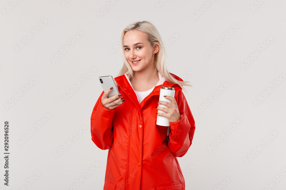 Pretty blond woman in a red raincoat holding white thermal mug with hot beverage and checking smartphone for notifications isolated over white background. Going for a walk on a rainy cold day Pretty blond woman in a red raincoat holding white thermal mug with hot beverage and checking smartphone for notifications isolated over white background. Going for a walk on a rainy cold day
