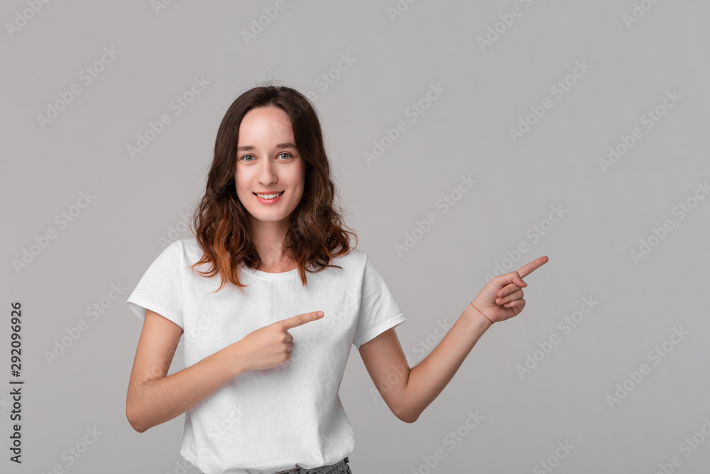 Pretty brunette woman in a white tee pointing at the empty space in a right side of a frame standing isolated over grey background. Copy space of advertising.