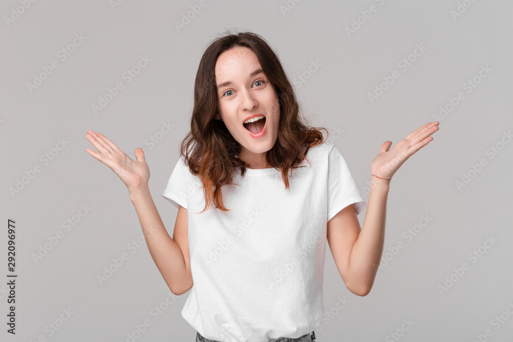 Excited brunette woman in a white tee shrugging her shoulders expressing emotion of surprise standing isolated over grey background. Emotion of surprise. Excited brunette woman in a white tee shrugging her shoulders expressing emotion of surprise standing isolated over grey background. Emotion of surprise.