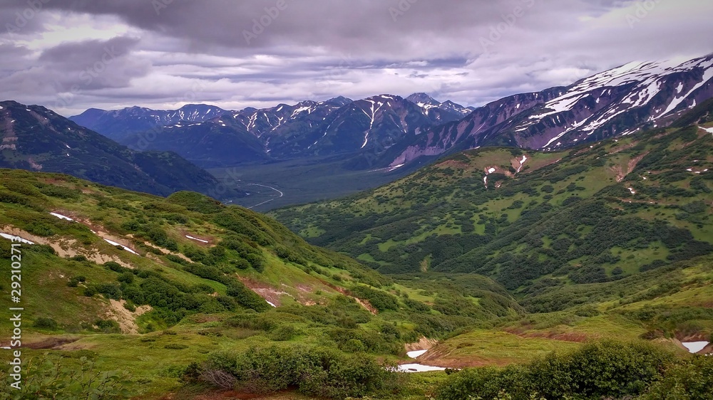 Fototapeta premium a mountain valley with a mountain road in the distance. Green fields, clouds.