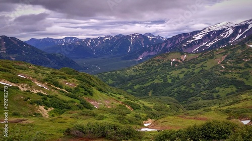 a mountain valley with a mountain road in the distance. Green fields, clouds.