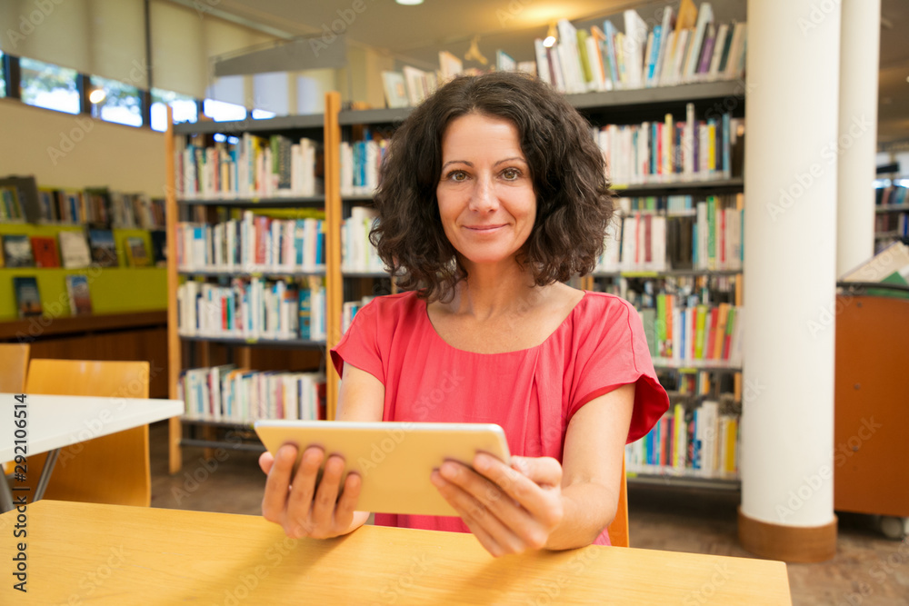 Satisfied female customer with gadget posing in public library. Middle aged woman sitting at desk with bookshelves in background, holding tablet, smiling at camera. Wireless connection concept Satisfied female customer with gadget posing in public library. Middle aged woman sitting at desk with bookshelves in background, holding tablet, smiling at camera. Wireless connection concept