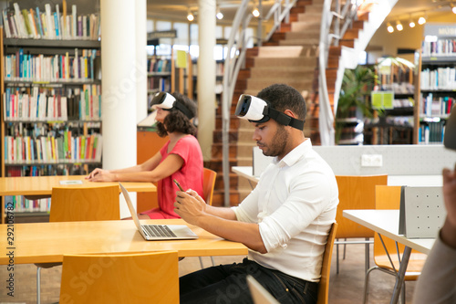 Adult students using VR technology for work on project in library. Man and woman wearing virtual reality glasses, sitting at desks, using laptop and smartphone. Innovation concept