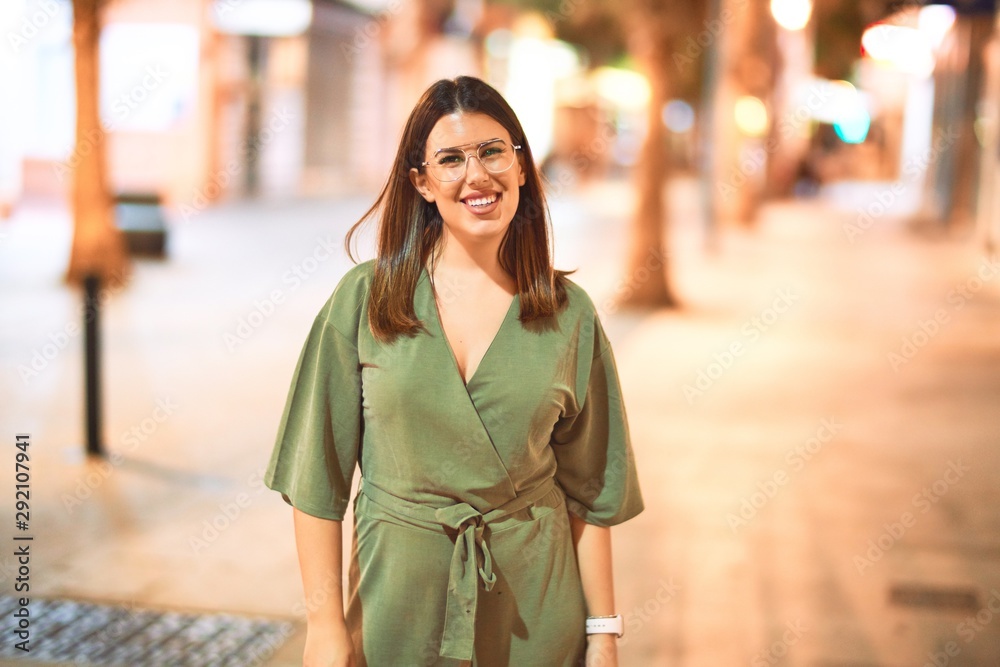 Fototapeta premium Young beautiful girl smiling happy and confident walking at the town street, standing with a smile on face around night lights bokeh