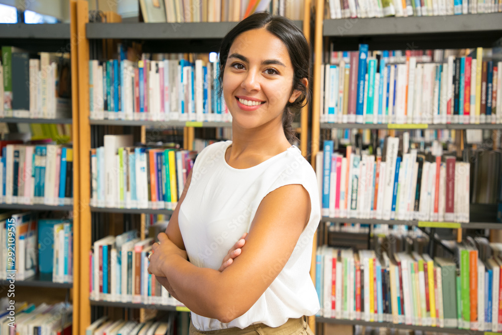 Smiling Asian woman posing at public library. Front view of smiling lady with crossed arms posing in front of bookshelves. Knowledge concept Smiling Asian woman posing at public library. Front view of smiling lady with crossed arms posing in front of bookshelves. Knowledge concept