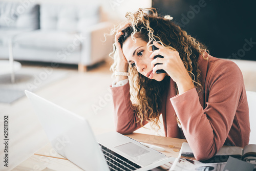 Cyrly busy business woman suffering stress working at laptop while talking on mobile phone in overwork. Tired businesswoman using phone. Girl feeling sad.