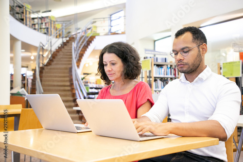 Focused college students taking online test in library. Man and woman in casual sitting at desk, using laptops, typing. Online exam concept © Mangostar
