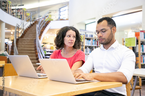 Adult female student consulting college mate in library. Man and woman in casual sitting at desk, using laptops and talking. Learning and communication concept