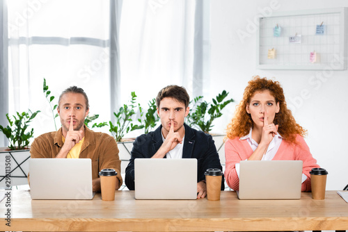 three friends sitting at table and showing shh gestures in office photo