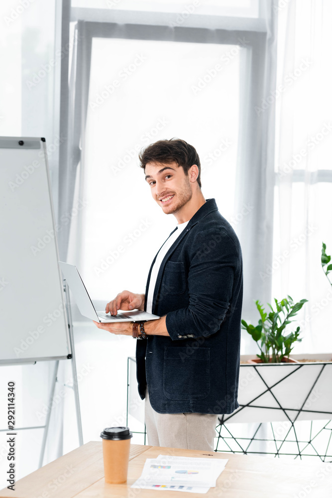 handsome and smiling man in shirt holding laptop and looking at camera in office