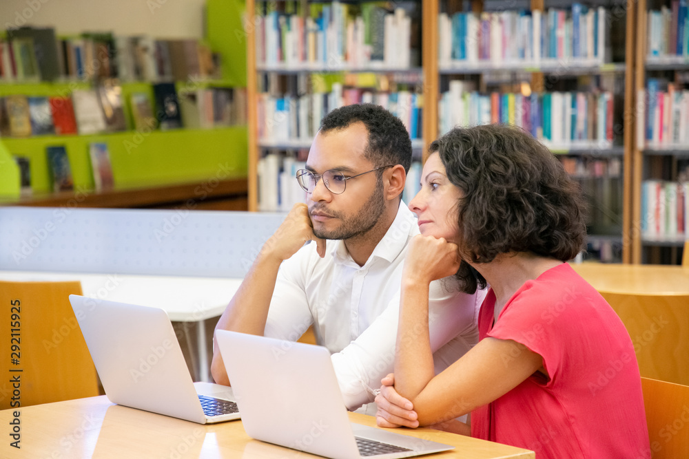 Pensive adult students watching content on computer in library. Man and woman in casual sitting at desk with laptops, thinking, looking at monitor. Video training concept