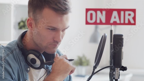 Tilt down of young Caucasian man sitting at desktop at laptop in broadcast studio and making notes on sheet of paper before broadcast photo