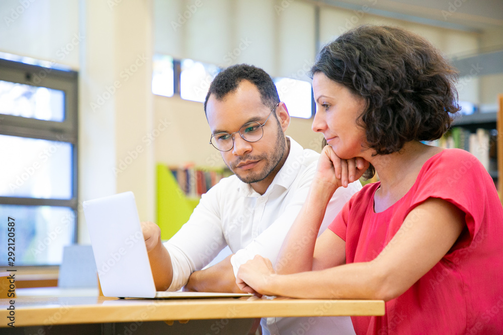 Two adult students working on class presentation in library. Man and woman in casual sitting at desk, using laptop and talking. Bookshelves in background. Exploring or research concept Two adult students working on class presentation in library. Man and woman in casual sitting at desk, using laptop and talking. Bookshelves in background. Exploring or research concept