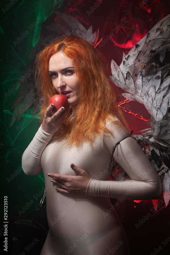 Fototapeta premium Mysterious plus size red hair girl in skincolor beige outfit with black and white big demon wings on gothic cemetery. Witch or dark angel style. Woman tempts by apple in the middle of the night