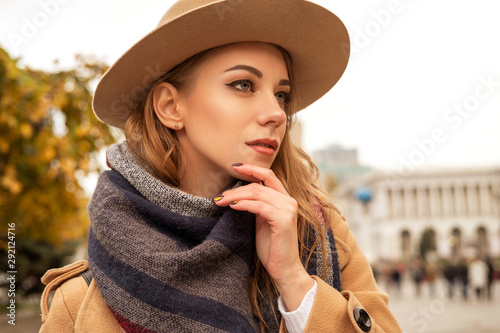 Blonde pretty woman on a walk in european old city center. Cold autumn weather. Girl wears brown coat, plaid scarf and stylish hat