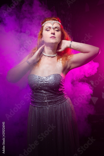 Mature plus size red hair woman in grey evening dress posing on dark smoke background ready for halloween.