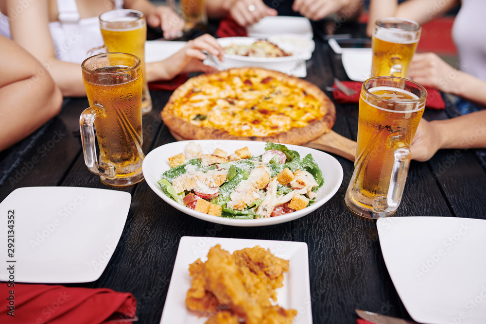 Close-up of salad pizza and glasses of beer are on the wooden table with young people sitting in the background