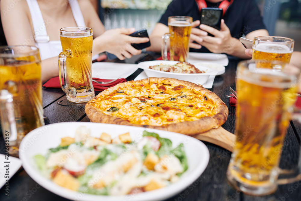 Close-up of young people sitting at wooden table eating pizza and salad and drinking beer at the restaurant