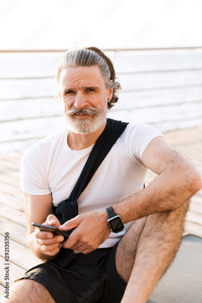 Image of satisfied old man typing on cellphone while sitting on mat Image of satisfied old man typing on cellphone while sitting on mat