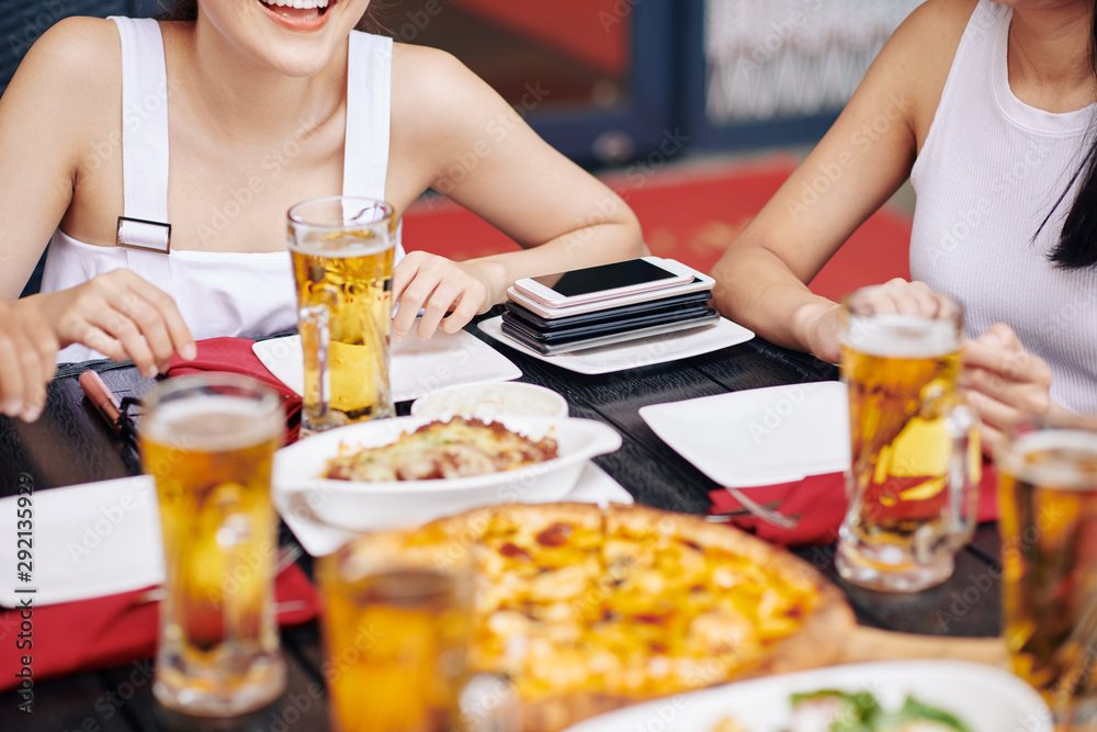 Close-up of young friends sitting at the table eating pizza and drinking beer with stack of mobile phones on the corner of the table Close-up of young friends sitting at the table eating pizza and drinking beer with stack of mobile phones on the corner of the table