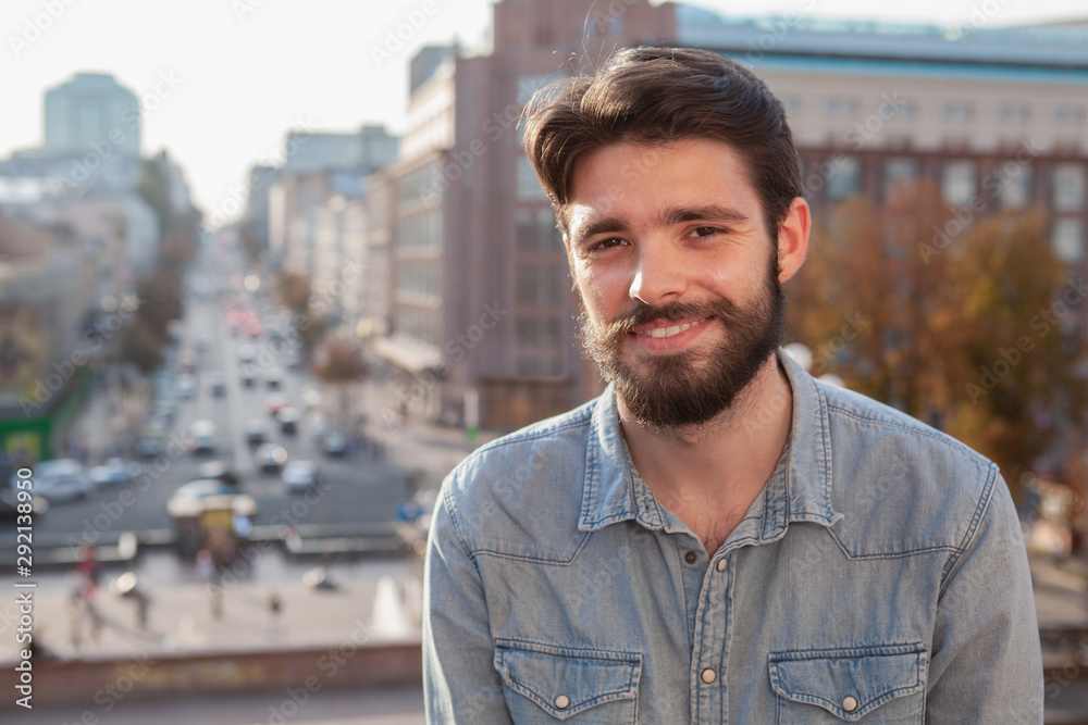 Happy handsome bearded man smiling to the camera, city streets on the background. Charming male traveler enjoying warm day outdoors in city center Happy handsome bearded man smiling to the camera, city streets on the background. Charming male traveler enjoying warm day outdoors in city center