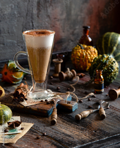 autumn traditional hot drink pumpkin spice latte in glass cup with foamy whipped cream on rustic wooden table with cinnamon, spoons and assorted mini pumpkins