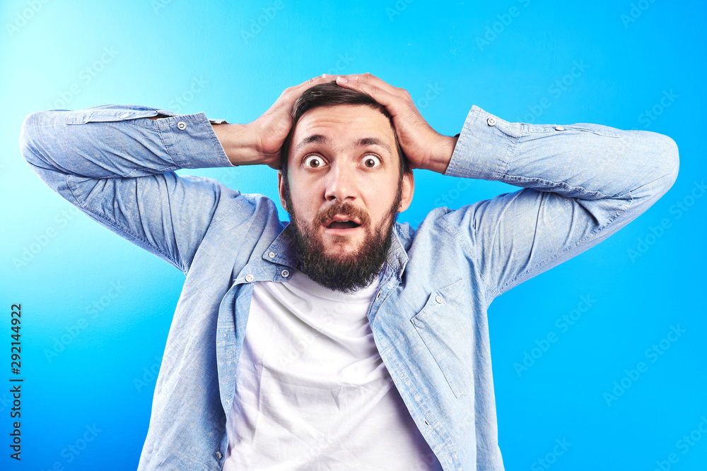 The excited face of the male buyer surprised at the price of the store. A man on a blue background looks at a very good deal. Grab your head with both hands The excited face of the male buyer surprised at the price of the store. A man on a blue background looks at a very good deal. Grab your head with both hands
