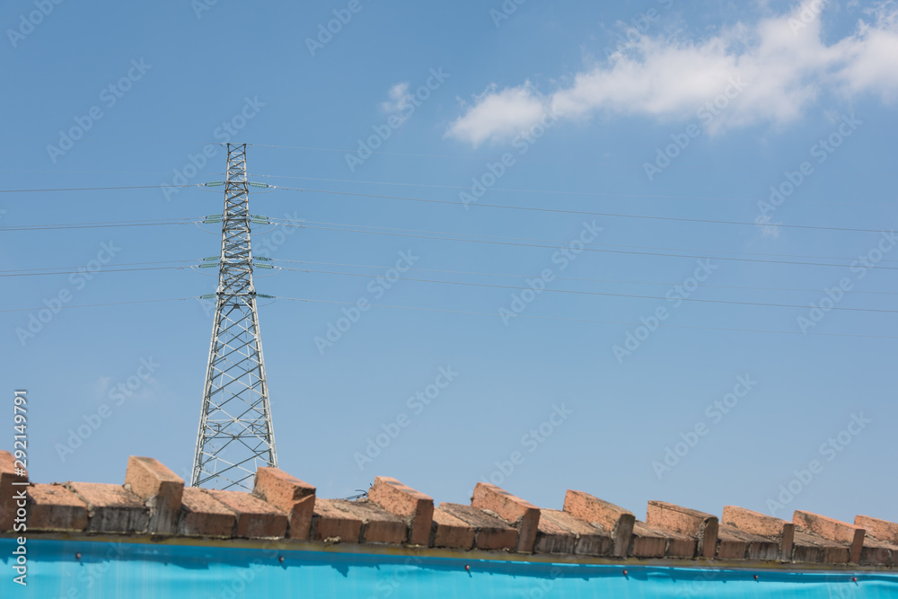 Outside the red brick wall, stands a high voltage electric tower on the background of blue sky Outside the red brick wall, stands a high voltage electric tower on the background of blue sky