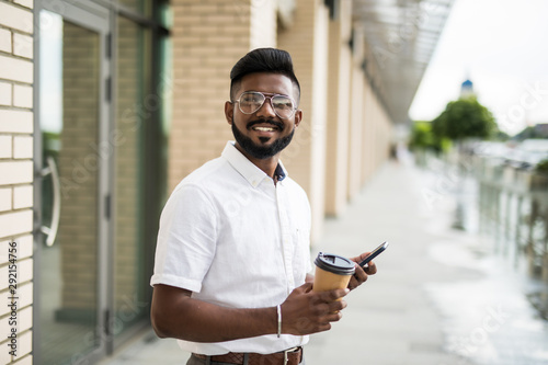 Smiling young indian man dressed in outfit and glasses resting outdoors enjoying leisure outdoors with coffee to go holding modern gadget connected to 5G wireless reading latest news on websites.
