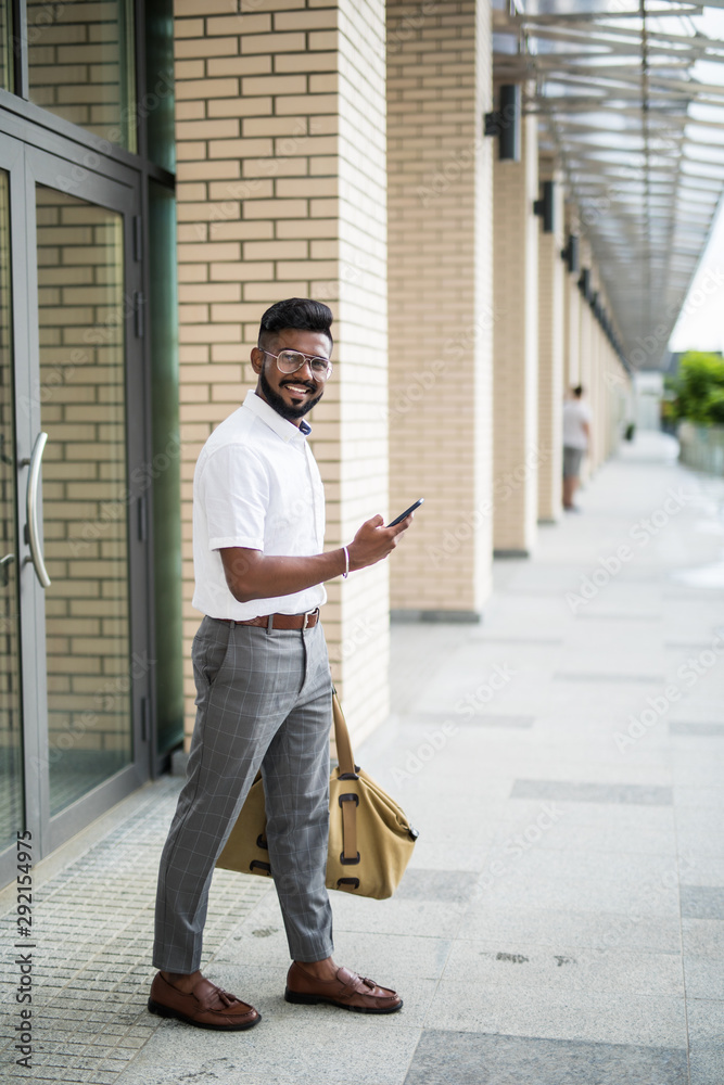 Young Indian Man with beard traveling carrying leather bag, walking on street talking on cell phone.