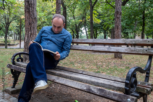 Middle Age Man Sitting on Wooden Bench in Park and Reading Book