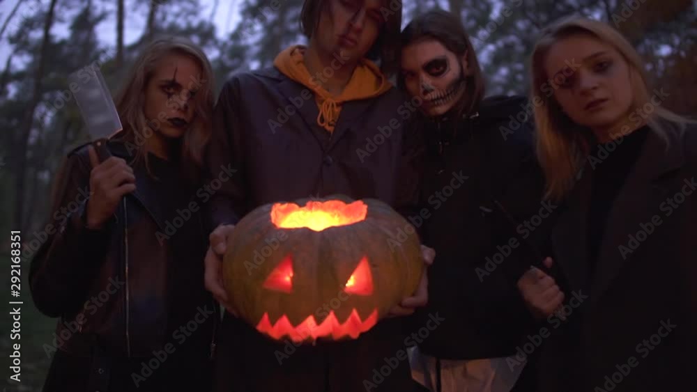 Tall handsome man holding halloween pumpkin with a light inside close up, three pretty girls standing near. People with scary makeup on faces looking into camera with evil looks. Bottom view Tall handsome man holding halloween pumpkin with a light inside close up, three pretty girls standing near. People with scary makeup on faces looking into camera with evil looks. Bottom view