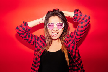 Rock'n'roll Kazakh girl in a plaid shirt on a red background playfully shows the tongue in glasses in the shape of hearts, studio portrait