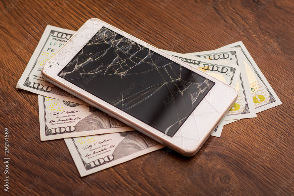 The broken phone lying on one hundred dollar bills, top view. Phone repair fee_ The broken phone lying on one hundred dollar bills, top view. Phone repair fee_