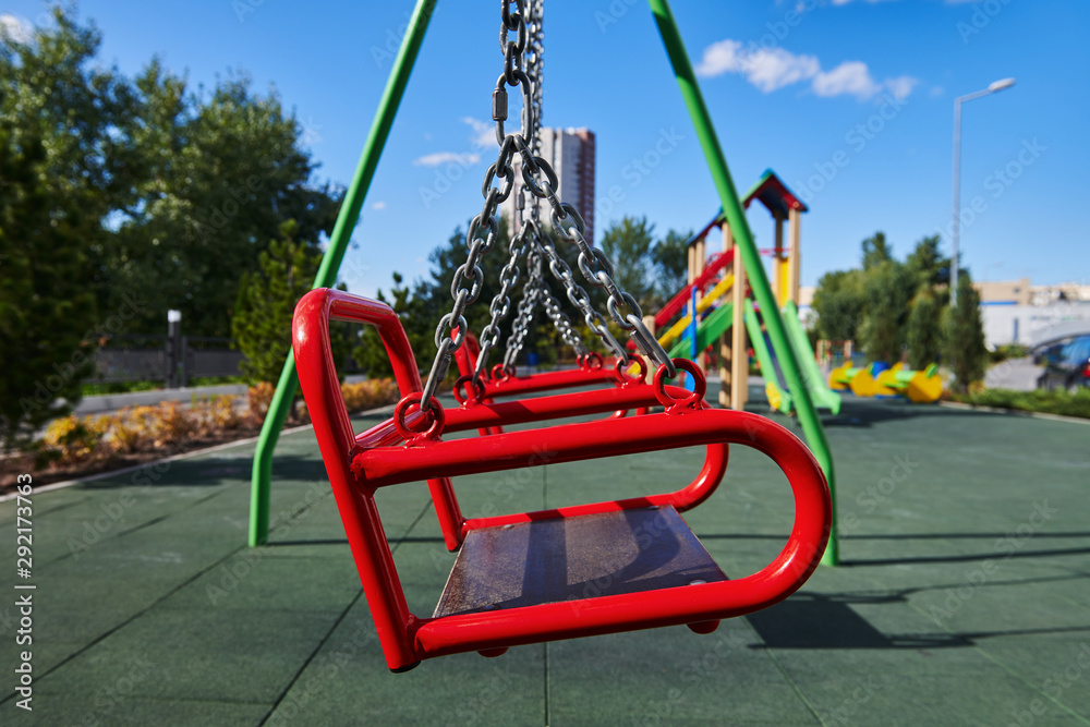Empty red swings on the playground against blue sky and trees.. Swing is hanging on the metal chains. A place for kids to have fun. Sunny day. Closeup.