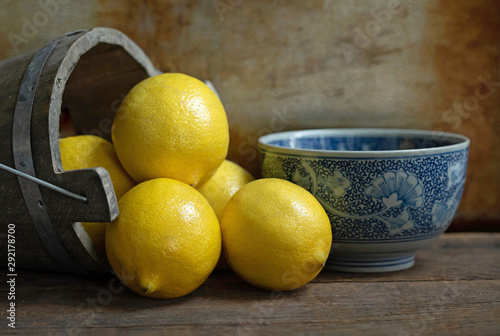 Original textured still life photograph of lemons pouring out of a wooden bucket with a blue china bowl on brown